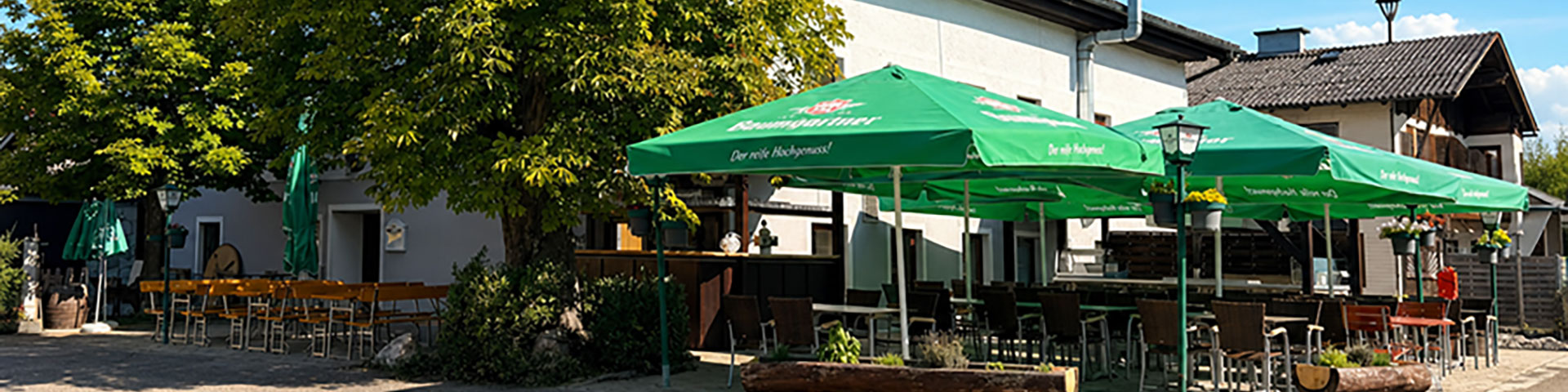 Wirt z'Hagenau - Fam. Probst | Außenansicht des Gasthofs an einem sonnigen Tag mit blauem Himmel. Große grüne Baumgartner Schirme überspannen den einladenden Gastgarten mit Tischen und Stühlen unter alten Bäumen.