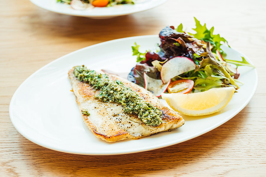 Wirt z'Hagenau - Fam. Probst | Gebratenes Fischfilet mit Kräuterpesto und gemischtem Blattsalat mit Radieschenscheiben und Kirschtomaten, garniert mit einer Zitronenspalte auf einem weißen ovalen Teller auf einem Holztisch.