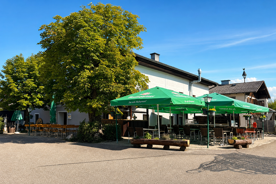 Wirt z'Hagenau - Fam. Probst | Außenansicht des Gasthofs an einem sonnigen Tag mit blauem Himmel. Große grüne Baumgartner Schirme überspannen den einladenden Gastgarten mit Tischen und Stühlen unter alten Bäumen.